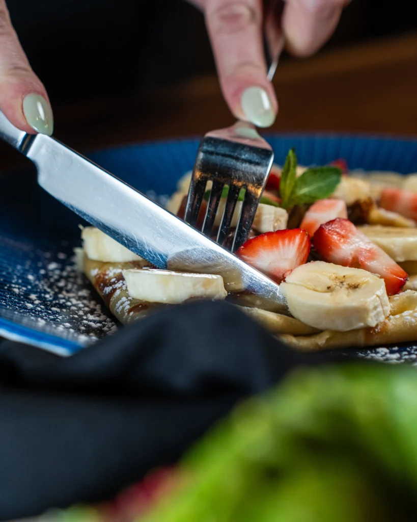 A knife and fork cutting into sweet crepes with strawberries and banana