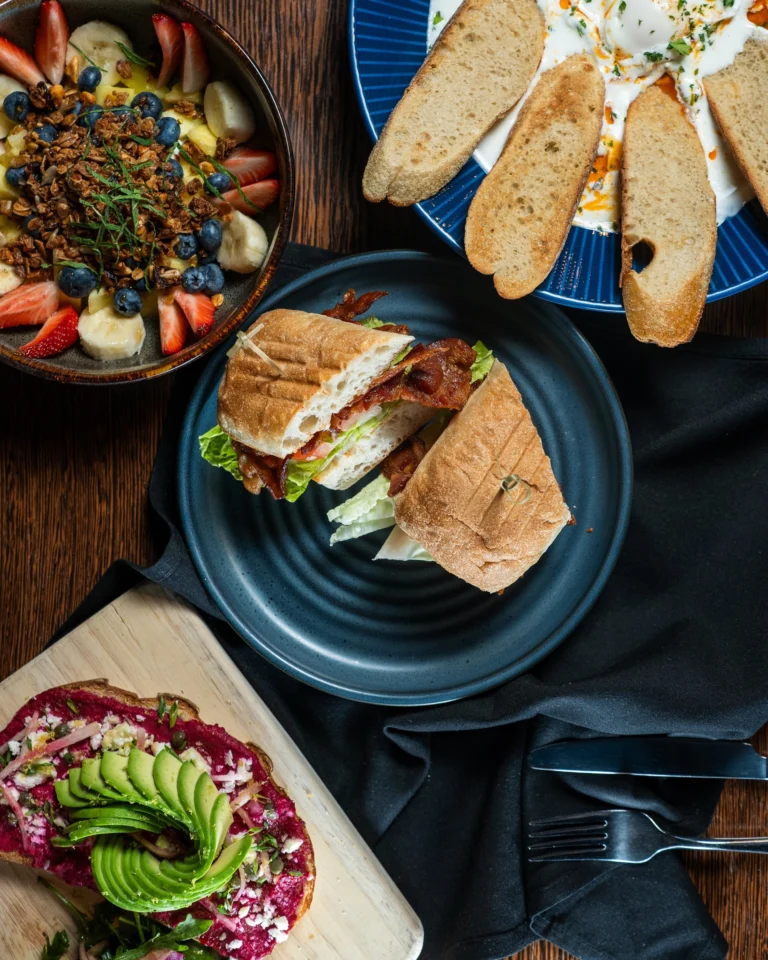 A stacked BLT on toasted bread beside a fruit and yogurt bowl