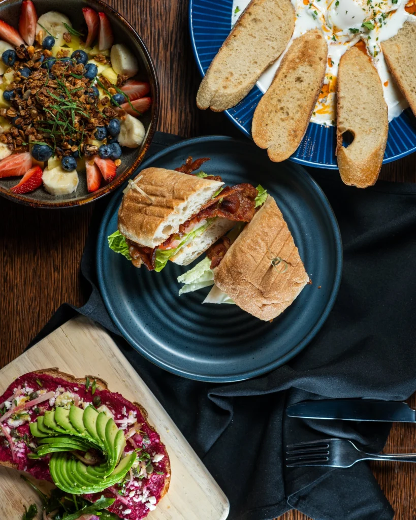 A stacked BLT on toasted bread beside a fruit and yogurt bowl